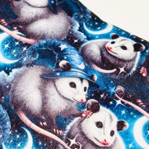 May include: A fabric with a repeating pattern of opossums wearing wizard hats, set against a starry night sky with crescent moons. The opossums are depicted in shades of gray and white, with pink noses and tails, on a blue background.
