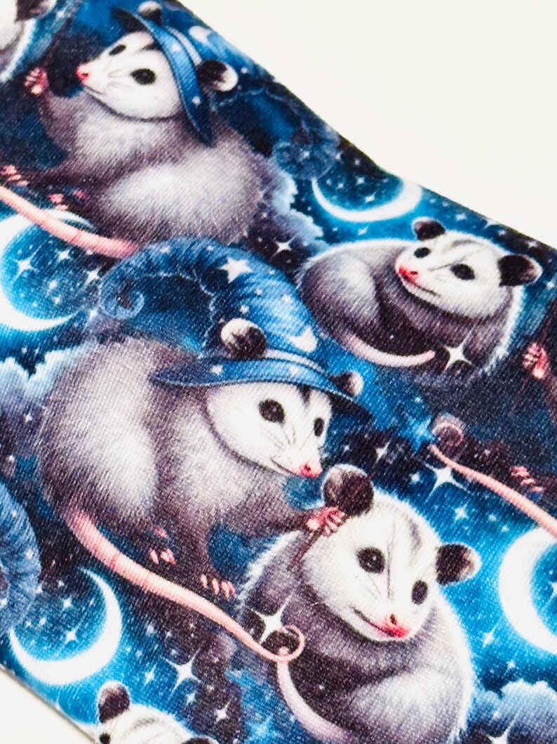 May include: A fabric with a repeating pattern of opossums wearing wizard hats, set against a starry night sky with crescent moons. The opossums are depicted in shades of gray and white, with pink noses and tails, on a blue background.