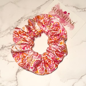 Puede incluir: Un scrunchie con estampado rosa y naranja sobre fondo blanco. El scrunchie tiene un diseño floral y abstracto. El scrunchie está sobre una superficie de mármol blanco. El texto "SUNSHINE scrunchies" está en una tarjeta rosa y blanca detrás del scrunchie.