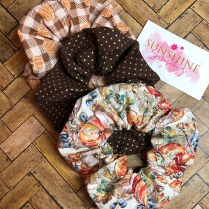 May include: Three fabric scrunchies in fall colors. One is tan with orange pumpkin pattern, another is brown with gold polka dots, and the third has a floral design with pumpkins and berries. A card with the text "SUNSHINE Scrunchies" is also visible.
