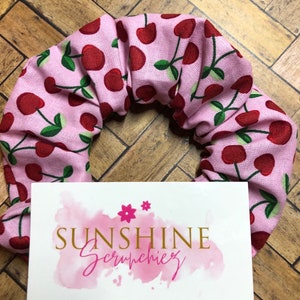Könnte beinhalten: Ein rosa Stoff-Haargummi mit einem Kirschmuster. Das Haargummi liegt auf einer Holzoberfläche mit einer weißen Karte, auf der "Sunshine Scrunchies" in goldenen Buchstaben mit rosa Aquarellhintergrund steht.
