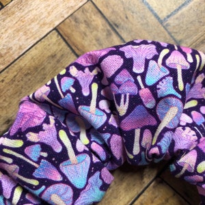 May include: Purple fabric scrunchie with a colorful mushroom print. The scrunchie is made of a soft, stretchy fabric and is perfect for holding hair in place.