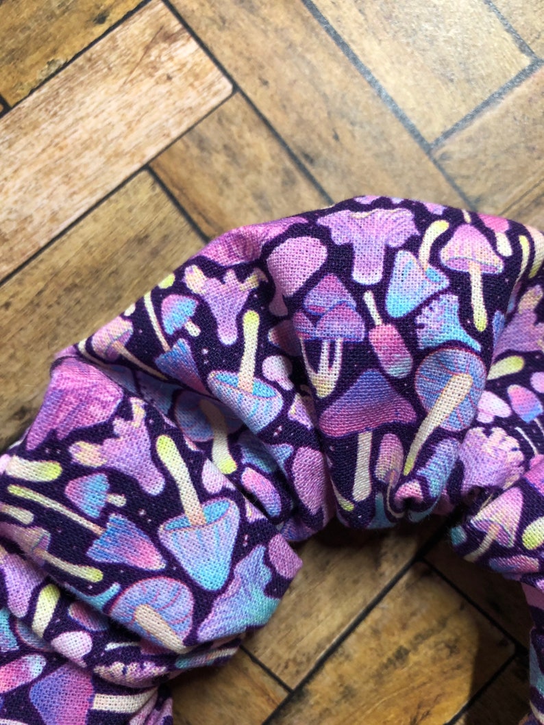 May include: Purple fabric scrunchie with a colorful mushroom print. The scrunchie is made of a soft, stretchy fabric and is perfect for holding hair in place.
