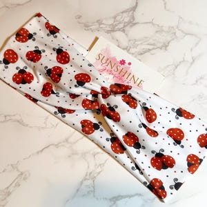 Peut inclure: Un bandeau blanc avec un imprimé de coccinelles rouges et noires. Le bandeau est torsadé et porte une petite étiquette qui dit "Sunshine Scrunchies".