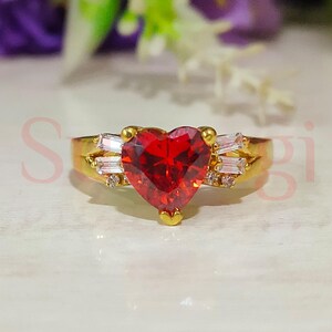 以下が含まれることがあります： 大きな赤いハート型の宝石と、バンドに小さな透明な宝石が付いた金の指輪です。