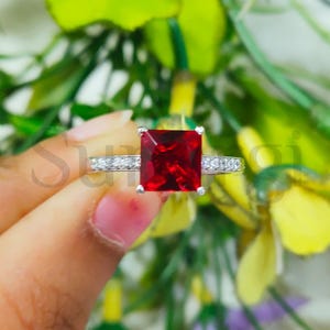 May include: A silver ring featuring a large, square-cut, vibrant red gemstone as the centerpiece. The band is adorned with smaller, clear gemstones. The ring is held against a backdrop of green and yellow foliage, highlighting the ring's design.