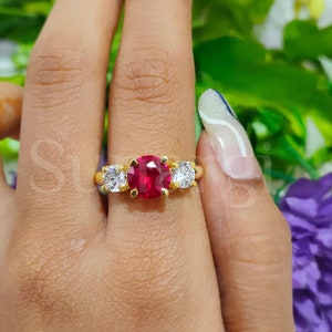 May include: A gold ring featuring a large, round, vibrant red gemstone flanked by two smaller, clear, round gemstones. The ring's band is a polished gold color, creating a classic and elegant design. The ring is set against a blurred background of green and purple.