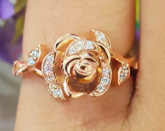 Puede incluir: Un anillo en forma de rosa con una banda dorada y piedras transparentes. La rosa es detallada y tiene un aspecto tridimensional.