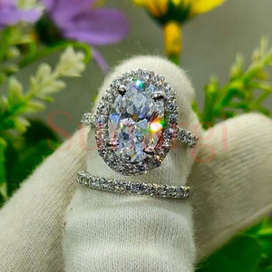 May include: A silver engagement ring set with a large oval-shaped centre stone surrounded by a halo of smaller stones. The band is also set with smaller stones.