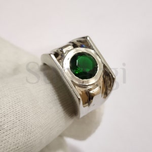 May include: A silver ring with a large, round, emerald-green gemstone set in a square bezel. The ring has a polished finish and a thick band. The gemstone is the focal point, set against a white background.
