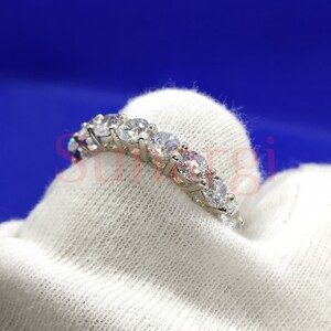 May include: A silver ring with a row of clear, round-cut gemstones. The ring is set on a white fabric against a blue background. The gemstones are evenly spaced and reflect light.