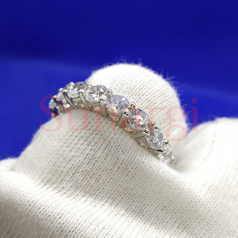 May include: A silver ring with a row of clear, round-cut gemstones. The ring is set on a white fabric against a blue background. The gemstones are evenly spaced and reflect light.