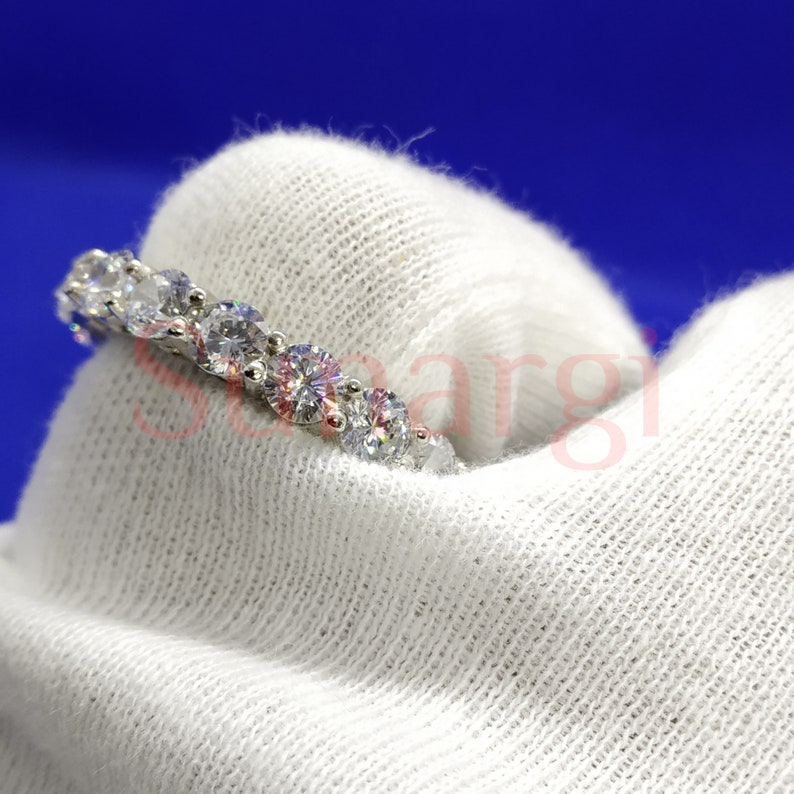 May include: A silver ring with a series of round, clear gemstones set in a row. The gemstones are evenly spaced and sparkle brightly. The ring is displayed on a white fabric background against a blue backdrop.