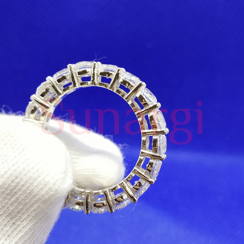 May include: A silver eternity ring with multiple square-cut, clear gemstones set around the band. The ring is displayed against a blue background and a white fabric.