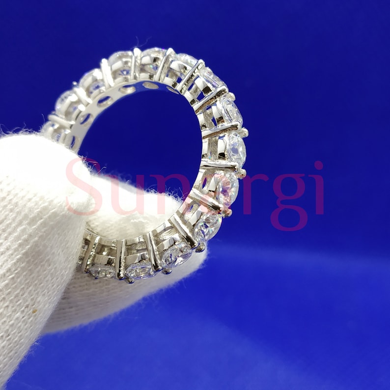 May include: A silver eternity ring with multiple round-cut, clear gemstones set closely together. The ring is held against a white fabric background, with a blue background. The ring's design allows light to pass through the gemstones.