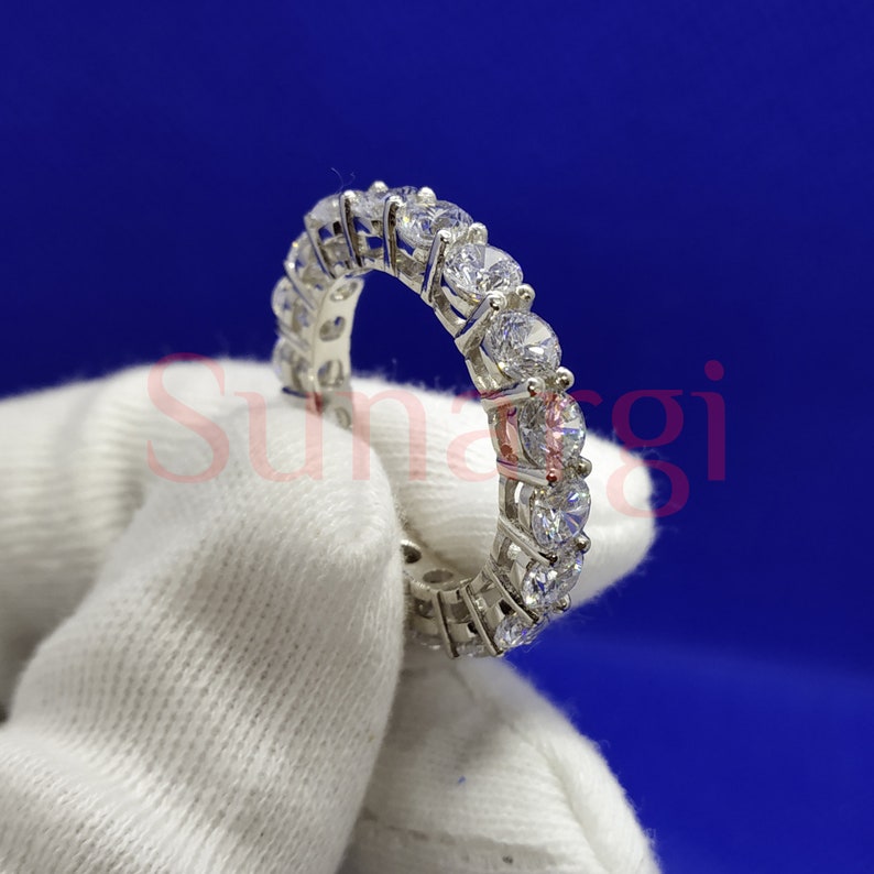May include: A silver eternity ring with a continuous row of round, clear gemstones. The ring is held against a white fabric background with a blue backdrop. The gemstones sparkle, reflecting light.