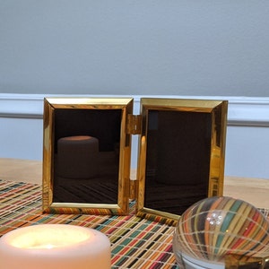 Puede incluir: Un marco de fotos doble dorado con dos marcos negros vacíos. El marco está sobre una mesa con una vela y un globo decorativo.