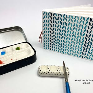 May include: A watercolour painting set featuring a black and silver tin, a white ceramic palette with red, blue, and green paint, and a patterned journal. A small ceramic brush rest with black dots and a paintbrush are also included.