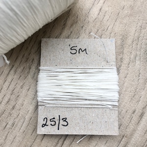May include: A spool of white thread on a cardboard label. The label says "5M" and "25/3".