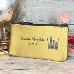 May include: Gold makeup bag with a zipper closure. The bag has the text "Trousse Maquillage Lucie" and a silhouette of makeup brushes printed on it.