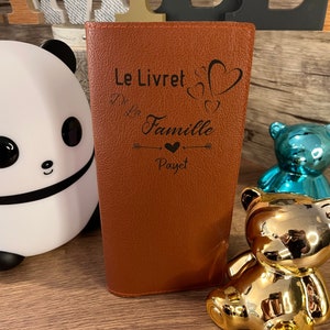 Peut inclure: Un carnet en cuir marron avec le texte "Le Livret De La Famille Payet" gravé sur la couverture. Le texte est entouré d'un motif de cœur et de flèches.