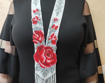 May include: A long, hand-beaded necklace featuring a white background with a detailed floral pattern of vibrant red roses and grey leaves. The design includes a pointed bottom edge finished with a fringe of white and light pink beads.