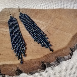 May include: A pair of long, dark blue beaded earrings with iridescent accents. The earrings are hanging from a wooden surface.