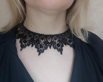 May include: A black beaded choker necklace with a scalloped edge. The necklace is made of small, dark beads and is worn around the neck. The person in the image is wearing a black top.