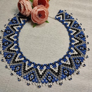 May include: A beaded necklace featuring a geometric pattern in shades of blue, white, and black. The necklace is crafted from small beads and has a fringe at the bottom.