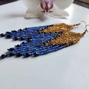 May include: A pair of long, beaded earrings with a gradient of blue, black, and gold beads. The earrings are dangle style and have a hook closure.