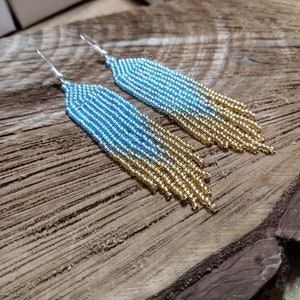 May include: A pair of long, beaded earrings with a gradient of blue and gold beads. The earrings are made with small, round beads and have a silver hook fastening.