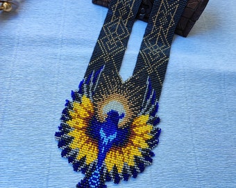 May include: A beaded necklace featuring a bird with outstretched wings in shades of blue, yellow, and black. The pendant is attached to a long, flat, woven beaded strap with a geometric pattern in black and gold, creating a detailed textile effect.
