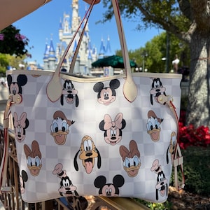 May include: A white tote bag with a checkered pattern and various Disney character faces, including Mickey Mouse, Minnie Mouse, Donald Duck, and Pluto. The bag has cream-colored handles with red trim and a cream-colored border.