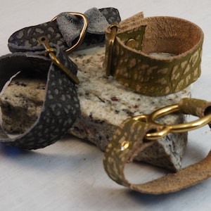 May include: Three leather bracelets with brass buckles. The bracelets are in shades of grey, green, and brown. The bracelets are on a grey and white speckled surface.