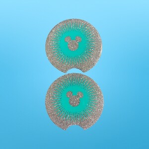 May include: Two round car coasters with a teal center featuring a Mickey Mouse silhouette. The coasters have a silver glitter design radiating outwards. The coasters have a small notch for easy removal and are set against a light blue background.