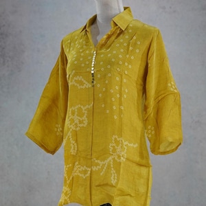 May include: A mustard yellow long-sleeve shirt featuring a white circular pattern and a vertical row of small metallic buttons along the front placket. The shirt has a classic collar and a lightweight, semi-sheer fabric with a subtle texture.