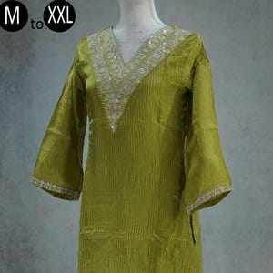 May include: Olive green two-piece outfit with a V-neck top and matching pants. The top features wide sleeves and silver embellishments around the neckline and hem. The pants have a loose fit. The outfit is displayed on a mannequin.