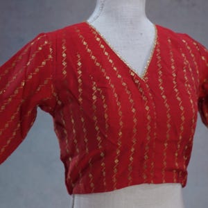 May include: Red wrap-style blouse with gold geometric pattern and elbow-length sleeves. The blouse has a V-neckline and gold trim along the edges. The fabric appears to be lightweight, suitable for warmer weather. The blouse is displayed on a mannequin.