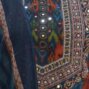 May include: A close-up of a colorful, patterned garment with a round neckline. The fabric features a mix of blue, orange, and green hues, with small reflective embellishments. A matching blue fabric is draped on the left side.
