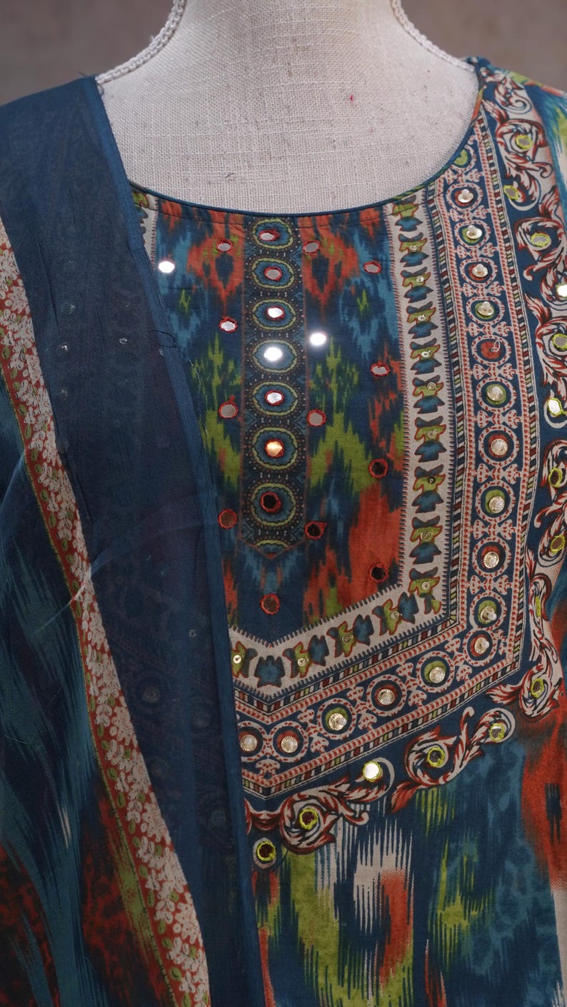 May include: A close-up of a colorful, patterned garment with a round neckline. The fabric features a mix of blue, orange, and green hues, with small reflective embellishments. A matching blue fabric is draped on the left side.
