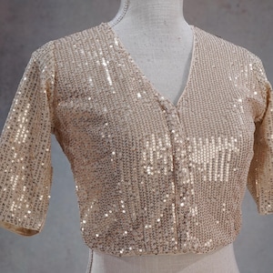 May include: A beige sequined top with a V-neckline and elbow-length sleeves. The top is covered in small, reflective sequins, creating a shimmering effect. The garment is displayed on a mannequin.