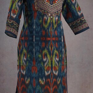May include: A long-sleeved, patterned tunic in shades of teal, orange, and green. The neckline and cuffs feature intricate embroidered details with small mirror accents. The tunic has a relaxed fit and a split hem.