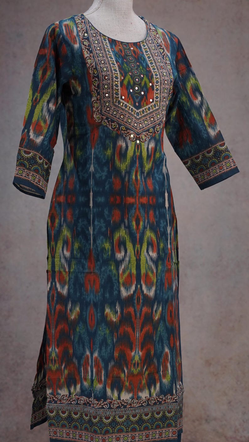 May include: A long-sleeved, patterned tunic in shades of teal, orange, and green. The neckline and cuffs feature intricate embroidered details with small mirror accents. The tunic has a relaxed fit and a split hem.