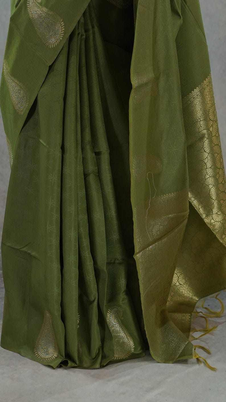 Puede incluir: Sari verde oliva con detalles dorados. La tela est&aacute; drapeada, mostrando patrones intrincados y un borde decorativo. El sari presenta dise&ntilde;os de cachemira y florales.