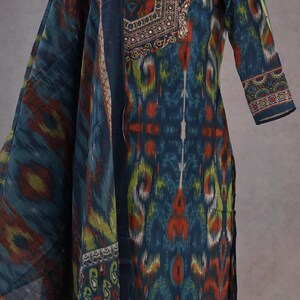 May include: A teal, orange, and green patterned salwar kameez with a matching dupatta. The tunic features a detailed neckline with small embellishments. The sleeves are three-quarter length, and the fabric has an ikat-style design.