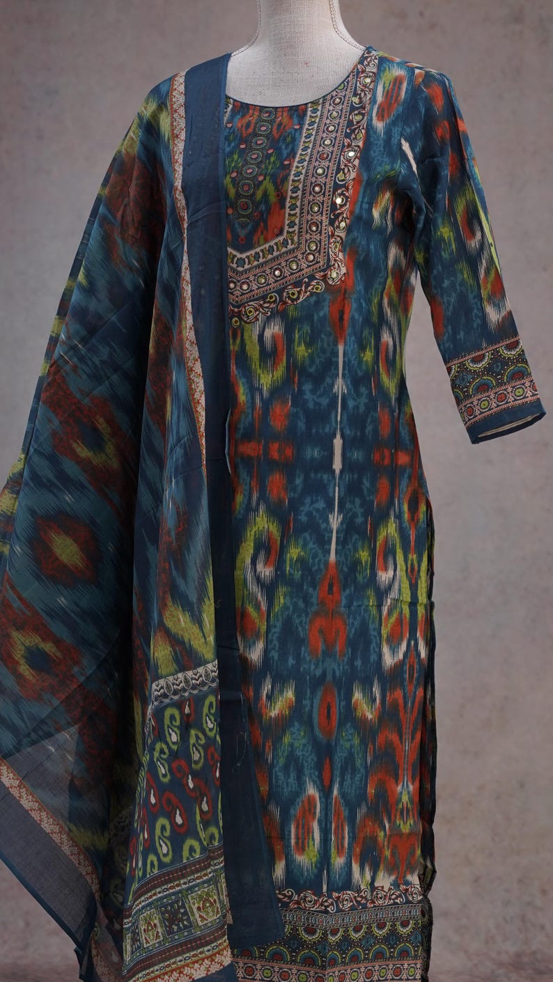 May include: A teal, orange, and green patterned salwar kameez with a matching dupatta. The tunic features a detailed neckline with small embellishments. The sleeves are three-quarter length, and the fabric has an ikat-style design.