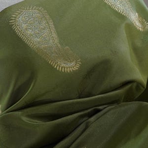 Puede incluir: Tela verde oliva con estampado de cachemir dorado. La tela est&aacute; drapeada, mostrando pliegues y sombras. El dise&ntilde;o de cachemir es detallado y se repite en la superficie, creando un textil lujoso.