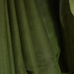 Puede incluir: Primer plano de tela verde oliva con sutiles patrones de estrellas. La tela est&aacute; drapeada en pliegues suaves, creando sombras y reflejos. La textura parece suave y ligeramente brillante, sugiriendo un material lujoso.