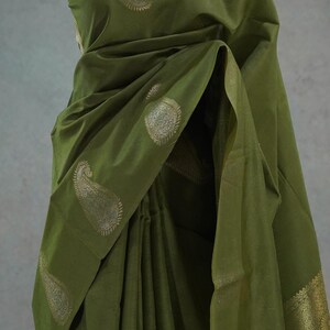 Puede incluir: Sari verde oliva con dise&ntilde;os de cachemira dorados. La tela est&aacute; drapeada sobre un maniqu&iacute;, mostrando los intrincados patrones y texturas. El borde del sari presenta adornos dorados detallados, agregando un toque de elegancia a la prenda tradicional.