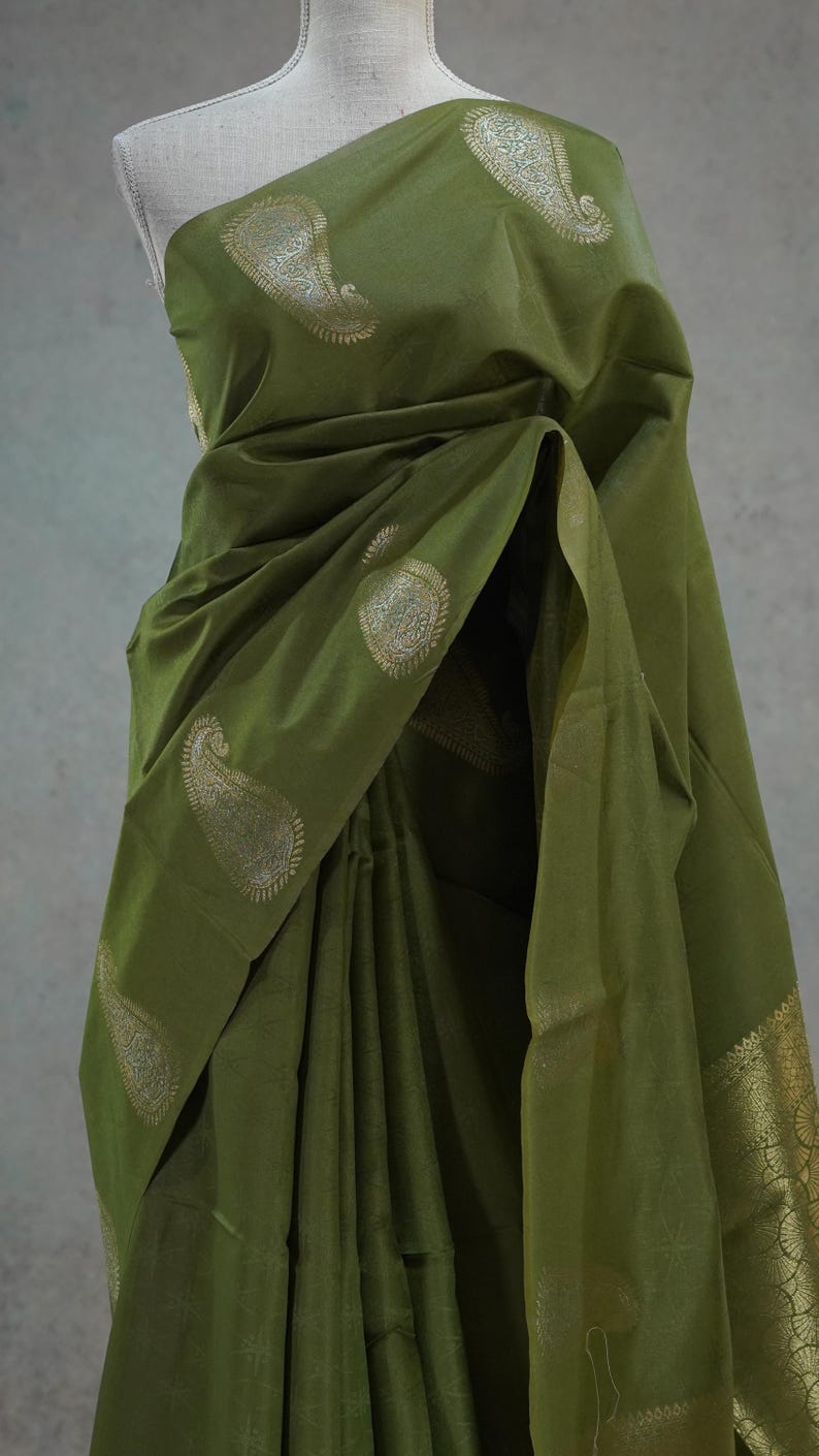 Puede incluir: Sari verde oliva con dise&ntilde;os de cachemira dorados. La tela est&aacute; drapeada sobre un maniqu&iacute;, mostrando los intrincados patrones y texturas. El borde del sari presenta adornos dorados detallados, agregando un toque de elegancia a la prenda tradicional.
